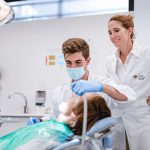 Un étudiant dentiste portant un masque et des gants examine la bouche d’un patient allongé sur un fauteuil dentaire, sous la supervision d’une professeure en blouse blanche, dans une salle de soins moderne.