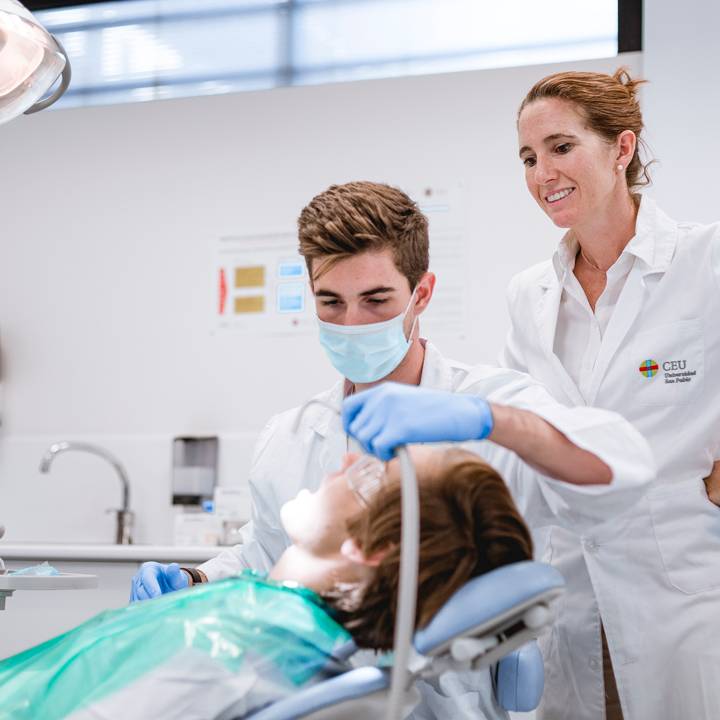 Un étudiant dentiste portant un masque et des gants examine la bouche d’un patient allongé sur un fauteuil dentaire, sous la supervision d’une professeure en blouse blanche, dans une salle de soins moderne.
